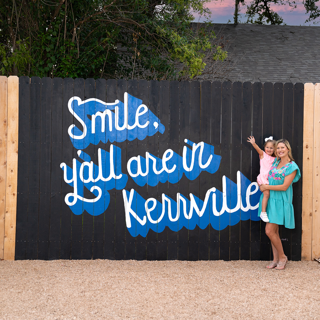 Woman holding her young daughter standing in front of a mural that says Smile You Are in Kerrville