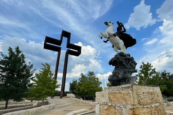 Large metal cross sculpture in the background behind a sculpture of a man on a horse