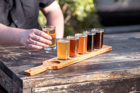 Flight of beer ranging from light to dark sitting on a wooden table with a man's hand reaching for a small glass