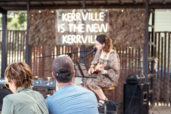 Woman playing guitar on stage outdoors in front of a sign that says Kerrville is the New Kerrville