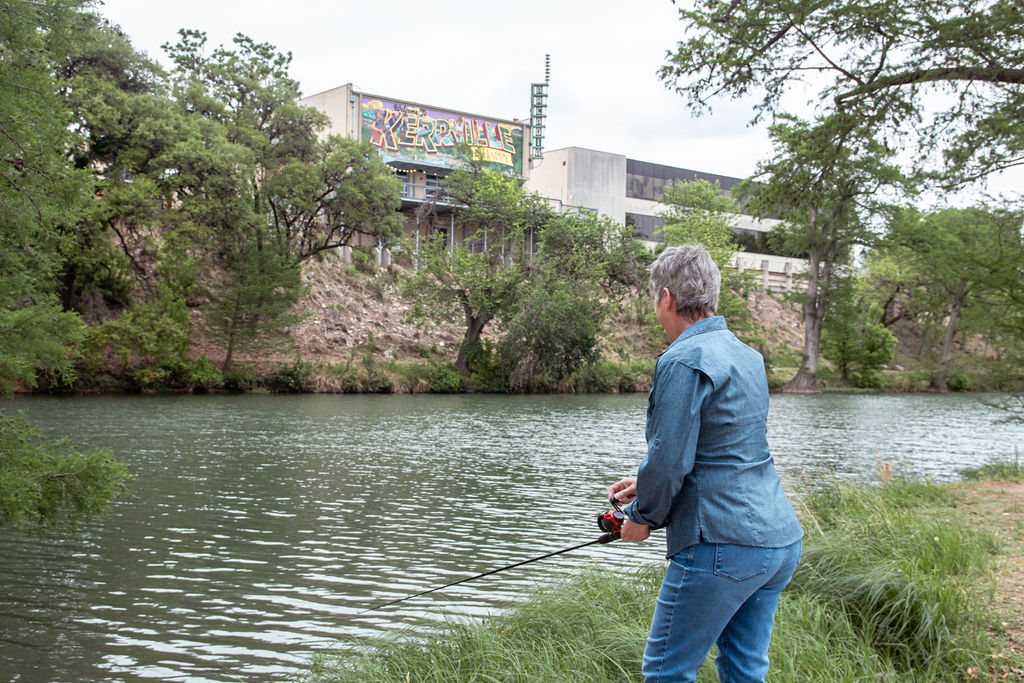 Woman fishing in Guadalupe River across from downtown Kerrville with mural on back wall of downtown building
