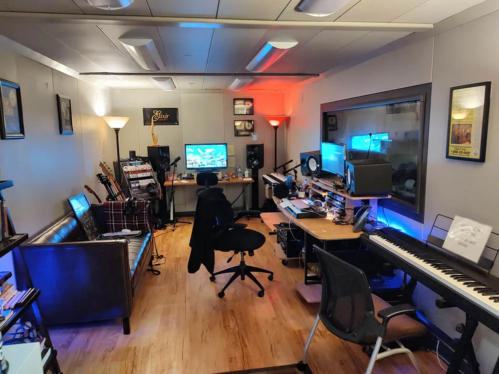 Small recording studio with wood floor and various computer screens, microphones, and instruments