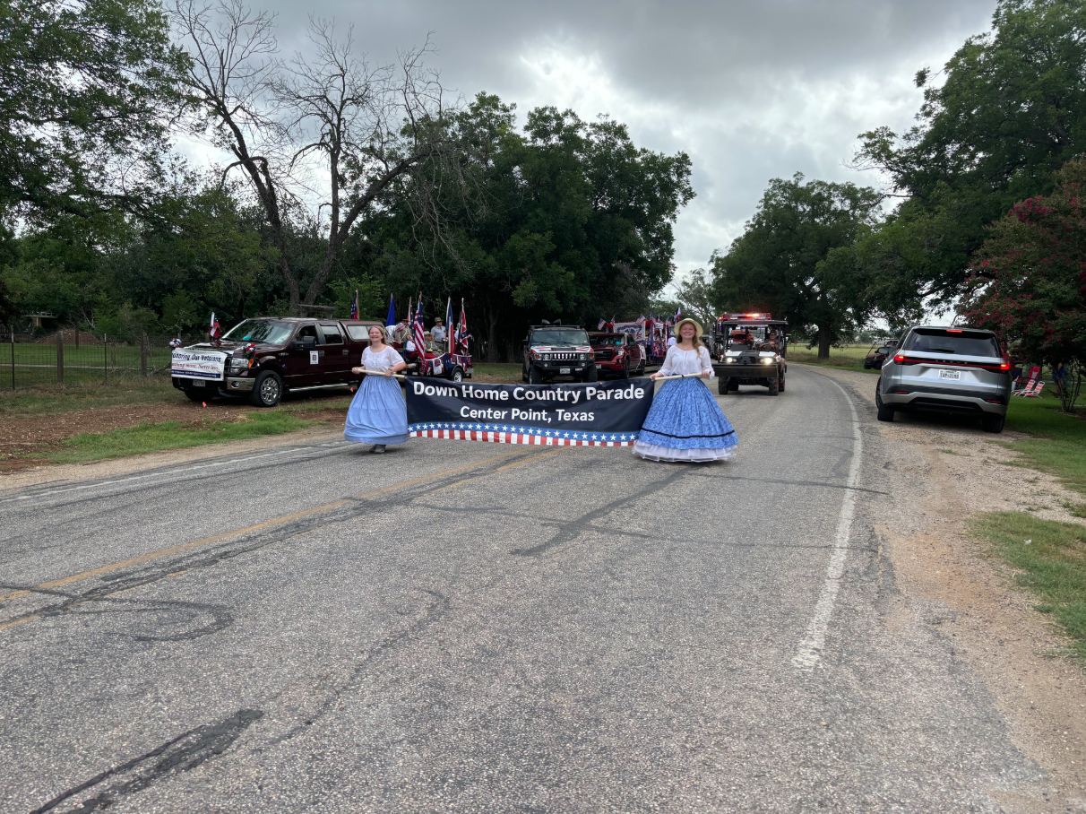 Women in blue dresses lead the Down Home Country Parade in Center Point, Texas, followed by trucks.