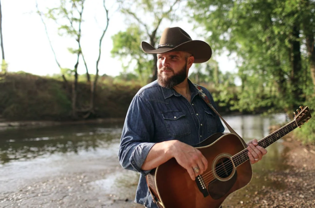 Promotional photo of musician Jake Hoot wearing a cowboy hat and holding an acoustic guitar by a river.