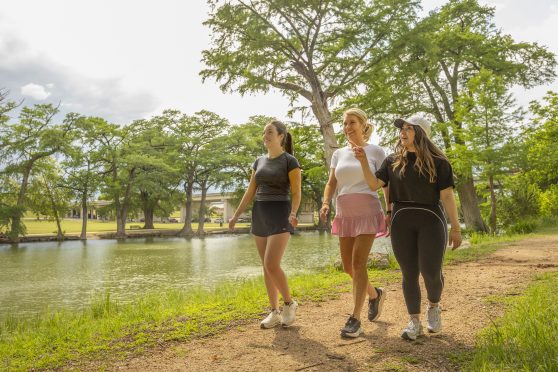 Three women smiling and walking along a grassy path next to a river with trees in the background.