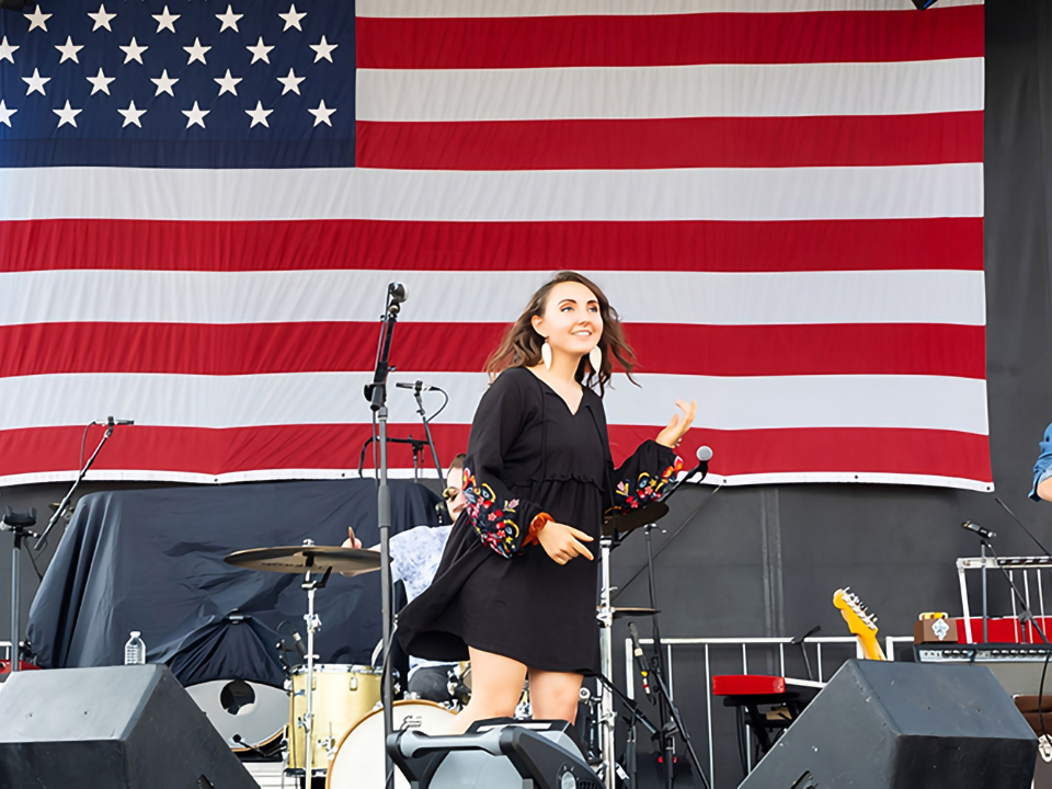 A woman in a black dress performing on an outdoor stage with musical instruments, set against a large American flag backdrop.