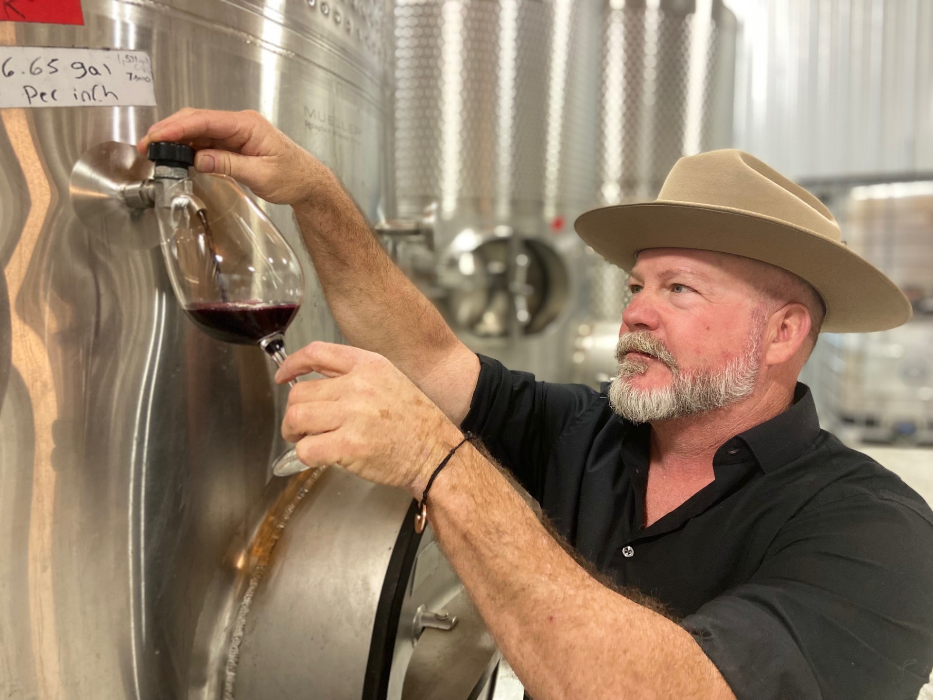 John Rivenburgh wearing a cowboy hat, pouring red wine into a glass from a large metal barrel.