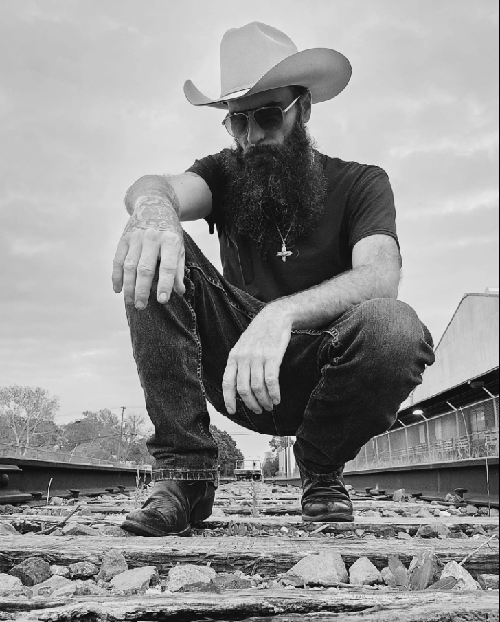 Black and white photo of musician Trick Myers wearing a cowboy hat and sunglasses on train tracks.